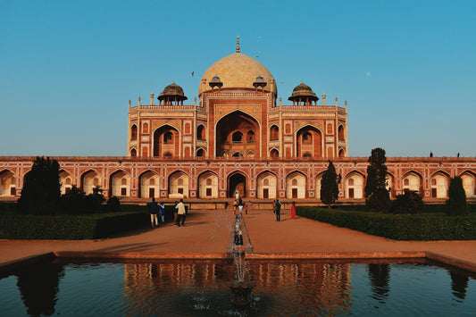 A large, symmetrical Moghul-era tomb (Humayun's Tomb) made of red sandstone with a central dome, reflected in a foreground pool and fountain.
