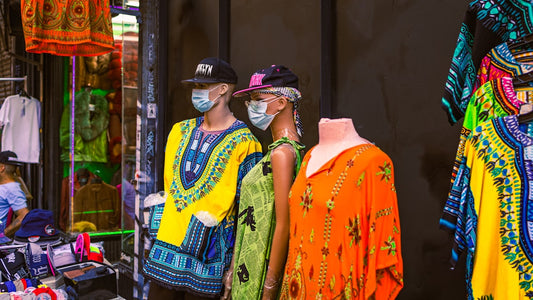 Three store mannequins wearing colorful, African-inspired dashiki shirts and dresses, hats, and protective face masks, displayed in a vibrant market or shop entrance.