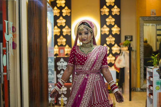 Bride wearing a traditional Indian bridal lehenga with intricate embroidery and gold jewelry, standing indoors with warm decorative lighting.