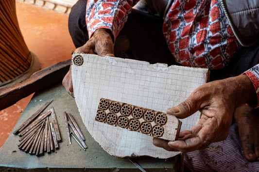Close-up of a craftsman's hands, wearing a red and white patterned shirt, holding a wooden block with a carved floral pattern, and resting it on a larger, marked piece of wood. Various carving tools lie nearb